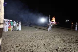 Night outdoor photo on a sandy open ground showing a ritual scene. On the left, a row of about six people in white and coloured clothes stand watching, including a figure in a green shirt and one in a red and yellow lungi. In the centre, a figure in white with a tall wooden pole stands near a lit smoky area. Behind them, an illuminated orange and red deity murti sits on a decorated platform with lights. On the right, a person in white holds a flaming torch beside another figure in dark clothing. Smoke drifts across the scene under a bright floodlight.