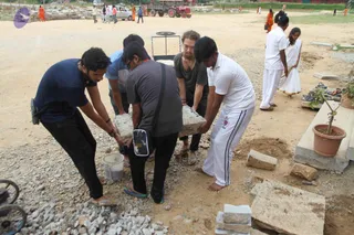 Outdoor photo at a dusty construction-type ground with gravel and loose stones. Five men bend together to lift a large grey stone slab, with a second slab on the ground. The man at left wears a navy t-shirt and dark trousers; the man in the centre wears a grey shirt and a crossbody bag; the man at right wears a white shirt with dark stripes and white trousers; a man in a blue shirt and a bearded man in grey top also help. Behind them, two people in white ritual clothing stand near a row of footwear and a potted plant, and an orange-clad person walks in the background beside a red tractor and parked vehicles.