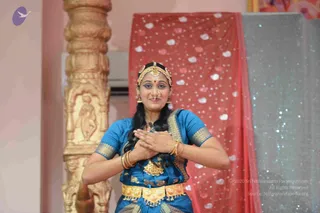 A female classical Indian dancer in a teal-blue silk costume with gold border and gold waist belt performs on a stage, hands pressed together at her chest in a namaskar-like gesture. She wears a gold head ornament, nose ring, drop earrings and a gold necklace. Behind her hangs a red curtain with white dot pattern and a sparkling silver backdrop, beside an ornately carved pillar. No a person candidate is visible.