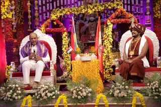 A festive stage decorated with garlands of marigolds and red flowers frames a seated gathering. On the right, a man in a rust-red turban, rust-red robe, white shawl and heavy gold necklace sits on a white and red throne, seemingly smiling. On the left, a bald man in a purple shawl and white clothes holds a microphone and speaks while seated on a matching throne. A framed portrait of a bearded saint in orange with rudraksha beads hangs behind them, flanked by saffron and red flags. Vases of white flowers and a golden draped table with flower arrangements sit in front. Two people are visible.
