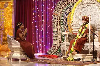 A woman in a rust-brown sari with jewelled earrings, a hair ornament and a beaded necklace sits on a silver ornate chair at left, hands pressed together in namaste while holding a microphone. Facing her on an elaborate silver throne with leopard-print cushions, a man in an orange-brown robe and matching turban wears a yellow and white flower garland and smiles, a microphone stand beside him. Behind them stands a large colourful jewelled backdrop with a red curtain and string lights, and at left a golden carved pillar. Stage setting for a question-and-answer satsang session.