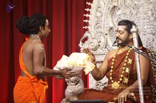 A photograph of a seated man in saffron-brown robes and heavy gold necklaces receiving a folded white and gold cloth from a standing attendant in orange clothing. The seated man wears a white forehead marking, a rudraksha-style mala and gold ornaments, and sits before an ornate silver throne with a peacock-motif carving, a microphone on a stand, a silver staff and a leopard-print cushion against a red curtain. The attendant, bare-shouldered in orange cloth with a hair knot and hoop earring, extends the offering with both hands.