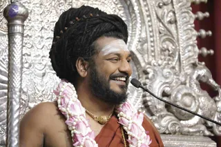 A smiling man sits before a microphone on a silver ornate throne with repoussé floral and figural carvings. He has long black hair wrapped up with rudraksha beads, a white vertical tilak on his forehead, and a full beard. He wears a pink lotus-flower garland with white beads over an orange-red saffron shawl and a gold chain pendant, and appears to be giving a talk. a person, seated in a teaching posture on a silver throne. Single person in a photograph.