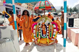 Outdoor procession scene at a Himalayan riverside town under a bright sky. In the centre, a long-haired man in saffron-orange robes, a rudraksha mala and a gold necklace, hands folded in greeting, stands beside a decorated palanquin carrying a deity murti of Venkateswara on a golden throne, framed by garlands of white, pink and yellow flowers. A red, green and gold umbrella shades the palanquin. Several other men in orange robes and orange caps stand nearby, one in a brown shirt on the right, with a speaker box and a blue-and-white striped pole in the foreground. Pale stone steps and houses fill the background. a person wearing saffron robes, rudraksha mala and gold necklace, leading the palanquin.