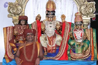A man in saffron-orange robes and a matching saffron turban sits in front of a raised blue dais, smiling, wearing a rudraksha-style beaded mala necklace. Behind him stand three large ornate deity figures with gold-and-red garments, jewelled necklaces, crowns with tilak marks, and golden arched backdrops. The central figure is a deity murti with a crown and a raised hand, flanked by two smaller figures in red-and-green and red robes. a person sits in front of the statues, wearing saffron robes, a turban and a mala, as the seated devotee presenting the murtis.