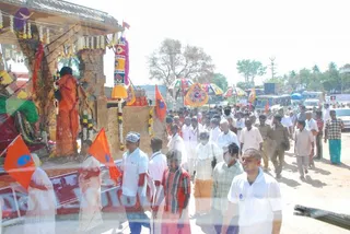 Outdoor procession on a dusty road with a decorated ratha chariot at left, its carved wooden pillars hung with flower garlands and a rear banner. Behind it, a crowd of men in white shirts and dhotis, some in checked shirts and sarongs, walks past parked buses and trees. Several orange flags and colourful umbrellas are carried above the marchers. A figure in an orange robe stands on the chariot beside a green-clad figure. A banner with a red and white design hangs from the chariot. Palm trees and power lines are visible in the background.
