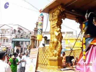 A photograph of a street procession with a gold-covered ratha (chariot) in the foreground. Its carved golden pillars show lion figures, and a decorated deity murti in a pink silk sari with white flower garlands stands at the right edge. Beside the chariot, a striped cylindrical banner with painted elephant and floral motifs hangs from a pole. Crowd members in white dhotis and saris, including a woman in a green sari, walk along the street, and a colourful temple gopuram with an arched gateway appears at left. A white placard with Tamil text is held in the lower left. A timestamp reads 12.03.2008 14:00.