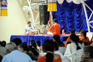 A indoor stage event with a blue draped backdrop and yellow fabric valances. On a blue-covered platform, a man in a light grey kurta sits at left playing a percussion setup with drums and a tabla-like instrument, while a performer in an orange robe plays a stringed instrument at the centre. Another person in orange sits at the right edge of the stage. Audience members sit in white plastic chairs in the foreground, many with long hair in braids, some in white and orange clothing. Water bottles sit on the stage, and white metal scaffolding frames a speaker.