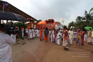 Outdoor procession on a dirt path in front of an orange-walled temple complex with a shaded pavilion and palm trees under overcast skies. Many devotees in white dhotis and shawls walk in a line, with several in saffron/orange robes, a woman in a patterned top carrying a long pole, and a man near the front holding a trident-topped staff. A man in white with a yellow shawl and a woman in orange sari carry umbrellas, including an orange umbrella and a green one. A cameraman in white stands at left filming near a black canopy, and a deity image is visible at the pavilion. Slippers are scattered on the ground.