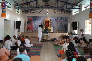 A seated speaker in orange robes and an orange head wrap sits on an ornate gold throne on a raised stage in a white-walled hall with ceiling fans. A large banner behind shows a peacock feather and a figure in orange, with Tamil text reading Bhagavat Katha and Dhyana Satsangam. Brass lamps flank the stage. A cameraman in white clothes stands beside a tripod camera in front of the stage. Around 30 seated devotees sit on striped mats, including shaved-headed men in white and orange, and women in white and red saris and salwar kameez. Two sound speakers stand on either side of the room.