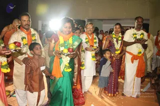 A photograph of a procession inside a hall with a white wall and doorway. A woman in a green saree with an orange shawl carries a silver plate of fruit (apples, bananas, oranges) and walks at the centre. Beside her, a smiling older man in white kurta and white dhoti wears an orange sash and marigold garland and holds a fruit plate. Other devotees in white dhoti or saffron-orange sashes, marigold and jasmine garlands, carry fruit trays. A young boy in a brown ethnic outfit walks holding a hand, and a small child in a blue shirt stands nearby. A shaved-headed man in saffron cloth and gold chain appears at the left edge. Strings of marigold garlands and flower petals decorate the scene, indicating a ritual offering.