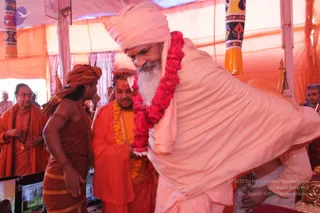A photo inside a pink-and-orange canopy tent at Kumbhapuri. A bearded man in a pale peach robe and matching peach turban, wearing a large red flower garland, bends forward. Beside him, a man in saffron-orange robes and orange turban stands with hands folded, wearing a yellow-orange garland. Behind them, a bare-chested man in orange cloth and an orange headwrap stands with arms at his sides, and a man in orange robes with a rudraksha mala gestures to the left. A ceremonial brass vessel sits at the lower right. Decorative fringed banners hang at the top corners.