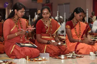 Three women in orange and red saris with gold borders sit cross-legged on a pale floor during a ritual. The woman on the left holds a small notebook and a pen beside a red container, the woman in the centre holds white cotton or flower pieces in her hands, and the woman on the right looks down at a phone. Each wears long rudraksha-style beaded malas, and the centre woman has a red bindi and white tilak. Brass pots, plates, small lamps and white offerings are arranged on the floor in front, and seated devotees in white and orange fill the background of a hall with white pillars. No a person candidate is visible.