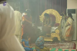 A smoky indoor ritual scene. A young man with long dark hair, wearing a rudraksha mala and ceremonial gold necklace over a saffron-orange garment, sits on an ornate gold throne with red cushions and a zebra-striped cloth. Two women in saffron and gold-bordered saris stand beside him; the woman on the right holds a flower garland and a lamp near the throne. A man in white kurta and a woman in a red sari stand behind, a child in blue sits on the floor, and an adult in a blue-and-gold sari kneels before the throne. Incense smoke fills the room. Background shows red curtains, a patterned rug and a white air conditioner. Possibly a darshan or puja setting.