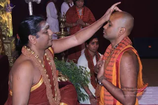 A seated man with long dark hair, a white tilak on his forehead and a multi-strand beaded gold-and-red necklace, raises his right hand to touch the forehead of a shaved-headed man in an orange-and-saffron shawl printed with script and a rudraksha mala. The recipient stands with hands folded at his chest and eyes closed. Behind them, a brass oil lamp, a potted green plant and a woman in a red shawl with hands folded stand in a hall with a gilded wall panel. A white-clad woman sits nearby. This appears to be a blessing or initiation moment during a samaya deeksha.