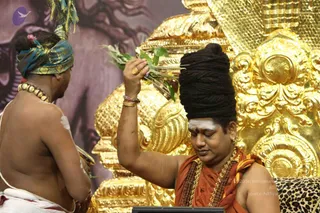 Photograph of a seated man with a tall, bound black hair crown and white sacred ash on his forehead, wearing an orange-red shawl and a rudraksha mala with ornate gold necklaces, holding a bundle of green grass stalks raised over his head. Behind him is an ornate gold throne with intricate carved floral and scrolled patterns. In the foreground a bare-chested priest with white ash markings on his back and shoulders, wearing a blue and gold turban and a beaded necklace, faces him and holds a brass vessel. The setting is a ceremonial stage with a purple-toned backdrop and a leopard-print cushion at lower right. Two physical people are visible. The figure in orange with the tall hair crown matches Swamiji (probable) by his crown, saffron clothing, rudraksha mala and gold necklaces.