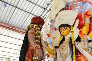 A photograph of a man in rust-orange clothing and a rust-orange turban, with a long dark braid hanging down his back, wearing a garland of white, yellow and red flowers and gold bangles on his arm. He stands with his head bowed and hands folded near a large deity murti of Devi with a yellow face, painted eyes, a white ornate crown, golden jewelry, yellow flower garlands and multiple arms. A red and colourful painted backdrop and a corrugated metal roof surround the scene.
