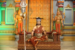 A seated man in saffron-orange robes sits cross-legged on a wooden throne draped with a leopard-print cloth, eyes closed in meditation. He wears a white headband with a crescent emblem, multiple rudraksha malas and a gold necklace, with bangles on his wrists. A tall silver trident stands beside the throne. Two painted deity statues with multiple arms, gold crowns and orange dhotis flank him in front of a large carved golden wooden door with ornate painted pillars. a person appears as the seated figure, with cues including the saffron robe, rudraksha malas, ceremonial gold necklace and throne.