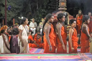 Night-time outdoor ceremony on a striped multicoloured mat with scattered yellow flowers. Four young men in saffron-orange dhoti-style wraps stand in a row with rudraksha malas and long dreadlocks, their bare chests marked with white ash lines. Behind them, more saffron-clad figures sit and stand near a reddish stone pillar with a garland. On the left, a woman in a white sari with a rudraksha mala and a child in a striped orange-and-white turban stand near seated spectators. Trees and lights frame the scene.