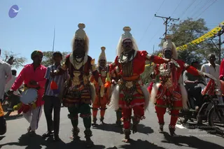 A street procession in daylight with several costumed dancers in tall white-and-gold headdresses, white plumes and bright red, orange and maroon costumes heavy with beaded necklaces, bells and ankle ornaments. Two dancers in front carry swords and wear green trousers and red garments; behind them a drummer in a pink shirt and green turban beats a drum, and a young boy in a striped shirt walks beside him. Spectators on motorcycles and roadside onlookers line the road under a clear sky, with a yellow police tape strip visible at right.