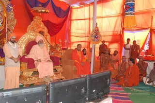 A photograph inside an orange and red tent at a religious gathering. An elderly white-bearded sadhu in a pale peach robe and orange turban sits on an ornate gold throne with red cushions, with a golden parasol above. Beside him a standing attendant in cream robes and an orange turban wears a white rudraksha-style mala. In front, a man in a saffron robe with a gold-bead necklace sits on a brown chair, flanked by seated monks in orange and ochre robes on a colourful striped mat. A small pennant with a yellow and blue emblem hangs from a pole, and two television screens sit in the foreground. a person is not clearly identifiable from appearance here, so the seated figures are described by role only.
