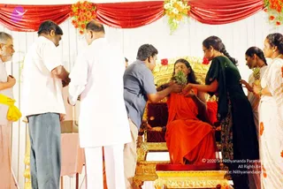 A seated figure in an orange-red robe sits on an ornate gold throne with a red cushioned seat, under a red draped canopy decorated with orange and white flowers. Several people stand around the throne offering devotion: a man in a blue shirt leans in holding green leaves toward the seated figure's head, a man in white shirt and trousers stands at left with hands joined, and women in dark green and white saris hold flowers and leaves nearby. One man at far left wears a yellow shawl. The scene resembles a ceremonial honouring at an outdoor stage. a person identity is not established from appearance, so the seated figure is described by role only.