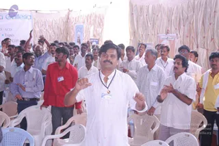 A photograph of a crowd standing among white plastic chairs under a pale draped tent at a person Spurana convention. In the foreground a man in a white kurta with a dark bead mala and a name badge raises his hands in front of him. Around him, men in white shirts, a red shirt, a yellow polo and checked shirts stand, some clapping and some with hands raised. A banner at upper left reads Galleria with the words DVD/VCD, Tapes and Gift. Portrait posters are visible on the tent walls. No identifiable a person figure is clearly visible in this image.