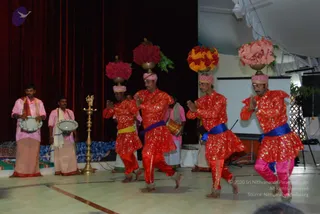 Four male dancers in matching red and gold costumes perform on a stage, each balancing a brass pot topped with red and orange flower arrangements on their heads. Their hands are folded in namaskar and they wear blue sashes and pink or red trousers with bells at the ankles. Two drummers in pink shirts and white dhotis stand at left playing hand drums, and a tall brass oil lamp with a bird-shaped top stands centre stage. A red curtain and projector screen form the backdrop.