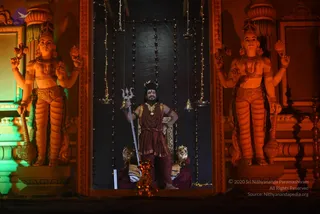 A man stands in a lit doorway-shaped shrine between two large orange-lit painted guardian statues with multiple arms and crowns. He holds a silver trident (trishul) upright in his right hand and rests his left hand on his hip. He wears a maroon-brown dhoti-style garment, a large gold ceremonial necklace, layered rudraksha malas, and a dark headdress with a small golden crown ornament. Behind him is a dark backdrop hung with string lights and small hanging bells, and a golden throne-like seat with lion figures is visible below him. Small brass oil lamps stand on the floor in front.