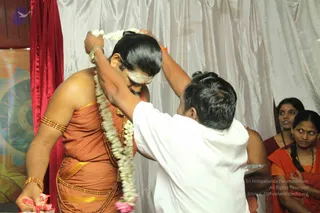 A photograph of a man in saffron-orange draped clothing with gold waistband and gold-bead armlets, having a long white jasmine garland placed over his head by a devotee in a white shirt. The recipient has a white vertical tilak on his forehead and his hair is swept back. Two women in orange saris stand behind, one with a gold necklace and a bindi, and a red curtain and white drapes form the backdrop. Pink flower petals and a small tray are visible in the lower frame. The recipient's appearance includes swept-back hair, gold ornaments and saffron clothing, with a devotee garlanding him, consistent with a person presence, though the identification is uncertain.