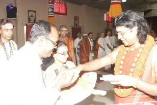 A photograph of a ceremonial moment indoors. A man with long swept-back dark hair, rimless glasses, rudraksha-style gold and beaded necklaces and an orange-saffron robe reaches forward and hands a small white item to a seated figure, while a man in white shirt and glasses and a woman in a white patterned top lean in. Behind them, several men in white dhotis with lanyards and orange-robed men stand along the wall; red and yellow hangings and framed pictures decorate the room. The seated figure in the foreground is a small child in yellow clothing lying across a white cloth. The orange-robed man with gold necklaces matches the page's Swamiji reference by appearance cues (long hair, ornate gold and bead necklaces, saffron robe), so he is labelled probable.