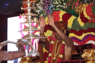 A seated man in an orange-brown turban and saffron-brown robe raises both palms in a blessing gesture before a flower-decorated seat banked with red, yellow and green garlands. He wears a long gold-bead necklace and a red and green garland with a bright multicolour band. On the left, a hand holds a tall silver multi-tiered aarti lamp with lit oil lamps, and a gold ornamental finial is visible at lower right. The background is a backdrop of colourful draping and white and orange flowers.