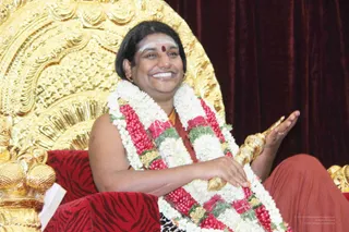 A smiling person sits on an ornate gold throne with a red velvet cushion and red tiger-striped fabric, against a deep maroon curtain. The figure wears a white and red flower garland with red and green accents, a white sacred ash mark with a red bindi on the forehead, and an orange-red garment. The right hand is raised in an open gesture while the left hand rests on a gold-tipped ceremonial staff. a person appears to be seated here, based on the swept-back hair, ceremonial garland and gold throne setting; the page describes a satsang welcoming visiting mahants and acharyas from Kashi Annapoorna Temple, Varanasi.