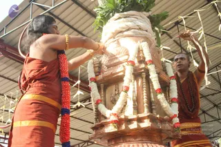 Outdoors under a corrugated metal roof, a long-haired figure in a rust-orange garment with a gold waist band and a bare upper body reaches toward a tall ornate copper or brass kalasha-like vessel wrapped with white cloth and topped with green leaves. Red and white flower garlands hang down its carved sides, and thin ribbon streamers are tied around it. A second man in a rust-orange robe with a rudraksha mala and a shaved head stands at right, holding up ribbons. Gold bangles adorn the first figure's arms. This appears to be a ritual decoration or abhishekam-style preparation of a sacred vessel.