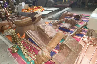 A workshop or storage area with copper and bronze ritual objects laid out on colourful striped woven mats. Large carved copper-toned pieces include a curved ornate base, a decorated seat or chair-like form, and a carved figure section with sculpted hands. Brass oil lamps with multiple tiers and cups sit on the left, a trident-like copper rod with tiered ends and a copper fork-like implement are on the right, and marigold garlands rest on a surface behind. No people are visible.