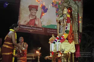 Night puja before a golden deity murti of a goddess, adorned with a gold crown, gold necklace and a long garland of yellow, white, red and multicoloured flowers, seated on an ornate golden throne-like arch. Two people stand at left: a woman with long hair in a rust-red and gold sari with a yellow flower garland, holding her hands over a brass oil lamp, and a shaved-headed man in orange-red dhoti with rudraksha mala and spectacles, appearing to chant. Brass oil lamps with flames sit in front of the deity. Behind them a large banner shows a smiling man in an orange turban and saffron robe with rudraksha mala, hands raised in blessing, against a world map with lit points; the banner reads Paramahamsa Nithyananda. The banner figure is a depiction; no clear appearance-based candidate is physically present.