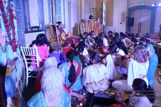 A large indoor hall with cream walls and a tall window, where a seated figure in a maroon-and-gold shawl sits on an ornate gold throne holding a long staff, facing a crowd of devotees seated on the floor. Behind the throne and a raised platform, an image of a deity murti is adorned with orange and white garlands. In the foreground, women in white and light blue saris and salwar kameez sit cross-legged with brass plates and oil lamps, and a painted backdrop of a blue deity figure stands at the left edge. A few men in white kurtas sit in the middle rows. Oil lamps and offerings are arranged on the floor.