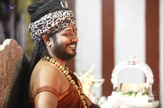 A smiling man in a leopard-print headwrap and rust-orange robe sits in a carved wooden chair on a stage. He wears a heavy gold bead necklace, a red rudraksha bracelet on his upper arm, and a black tilak on his forehead, with long dreadlocked hair falling over one shoulder. Beside him are a silver kalasha-style vessel and a silver arched shrine or peetha with flowers. Background pillars are red and white. One person is visible.