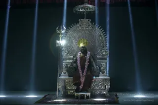 A seated figure on an ornate silver throne with a silver canopy and carved lion-shaped armrests sits centrally on a patterned rug before a dark backdrop lit by vertical blue light beams. The figure wears a gold crown-like headdress, a pink and white flower garland, and an orange-brown robe, and holds a silver trident. The figure's appearance matches the presentation cues of the candidate named on the page, including the crown, saffron-toned clothing and garland, but the face and features are not clearly discernible, and the figure could also be a statue or a depiction, so identity is uncertain. A copyright credit overlay appears at the lower right. No other people are visible.