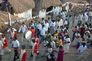Elevated view of a village street lined with thatched-roof huts and strings of small blue-and-white pennant flags. A procession moves down the dirt road: a brass band in red and white uniforms with peaked caps, playing a large drum, cymbals and tubas, leads the way. Behind them, many people in white shirts, saris and salwar kameez walk in a wide group, several women carrying silver trays or pots with flower offerings, while onlookers stand near the huts and a few children appear in the foreground. No single figure can be identified as a person with confidence, so the presence of any such person is uncertain.