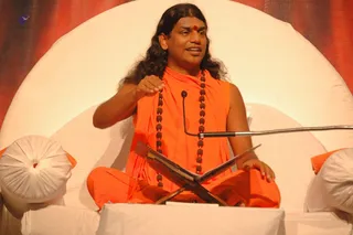 A seated man in saffron-orange robes sits cross-legged on a large white circular cushioned seat against a red-and-orange draped backdrop. He wears long dark swept-back hair, a red tilak on his forehead, and multiple dark rudraksha mala beads around his neck. His right hand is raised in a gesture while he speaks into a gooseneck microphone. A wooden book stand holds an open book on a white-draped table in front. a person is shown in an appearance-based label, giving a discourse at the event.