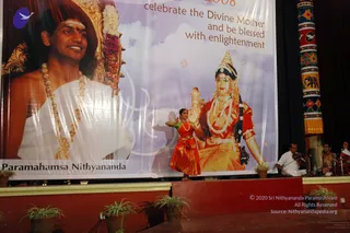 A stage at an evening cultural programme with a large white banner behind it. The banner shows a smiling man with shaved head, forehead tilak and a gold ornate necklace over white robes, labelled Paramahamsa Nithyananda, beside a painted goddess figure in red and gold with a crown and garlands, and text reading celebrate the Divine Mother and be blessed with enlightenment. On stage a dancer in a red and orange costume performs in front of the banner. At the right edge, a seated musician in white clothes plays a violin, with two seated singers or musicians beside him. Potted plants line the stage edge and a decorative painted pillar stands at the right.