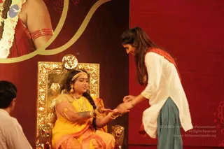 A stage photograph from a KAILASA event. A woman in a mustard-yellow sari with gold jewelry, bangles, a maang tikka and a long braid sits on an ornate gold throne carved with lion heads, a gold sun-disc behind her. She extends her hands to a younger woman in a white kurta with teal trousers and a red-orange shawl, who bends forward to take them. A man in white clothing is partly visible at the lower left. The backdrop is deep red with a gold scroll motif and a garlanded figure with red armlets at the top left. This is a staged performance or ritual demonstration rather than a seated darshan.