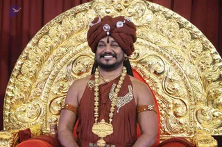 A smiling man seated on an ornate gold throne with a large carved back and red curtain behind him. He wears a rust-red turban with a silver crescent and jeweled ornament, a red-orange cloth wrap over his shoulders, a long gold necklace of beads with a large gold pendant, a gold collar piece, and red beaded armlets. Probable Swamiji by appearance cues: turban, ornate gold and rudraksha-style adornments, and the throne setting. A small brass vessel sits near the throne. Single person visible.