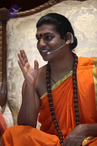 Close photo of a smiling man seated on an ornate cream and gold upholstered throne, raising one hand in a gesture while speaking. He wears an orange saffron shawl with a gold-bordered edge, a long multi-strand rudraksha mala with silver spacers, and a red tilak on his forehead. A small headset microphone is attached near his mouth and a earpiece is in his ear. Hair is swept back into a bun. Swamiji (probable) is a seated speaker in saffron with rudraksha mala and a swept-back hair bun, presented as a discourse speaker.