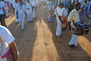 A dusty dirt road in warm low sunlight, with long shadows cast by a crowd of men in white. In the foreground, a man in a cream shirt and white dhoti plays a double-headed drum slung from his shoulder, with a blue cloth and red-and-white strap. Beside him, another drummer in white with a yellow shawl and orange sash plays a small drum. Behind them, many men in white dhotis and shirts, some with orange and blue scarves, walk along the road, and a man in a light blue shirt carries a lanyard badge. The scene suggests a drum-led procession on the road.