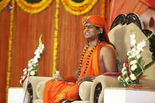 A man in saffron-orange robes and an orange turban with a jewelled brooch sits in an ornate dark-wood chair on a stage, speaking into a headset microphone. He wears long rudraksha mala beads and has a red tilak on his forehead; his hands rest on the chair arms. Behind him hang orange drapes and yellow marigold garlands, with white and red flower arrangements beside the chair and a water bottle on the floor. A partial printed portrait appears at the upper right.