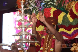A photograph of a person in rust-orange robes and a matching orange turban, with both palms raised in front of them in an addressing or blessing gesture. They wear a multi-strand garland of red and yellow flowers over the shoulders, a gold necklace, and gold armlets. Behind them is a large arrangement of red, yellow, white and purple flowers, and at left a tall silver multi-tiered lamp with lit flames stands before a colourful backdrop. The scene reads as a dais or stage setting at a devotional gathering. One physical person is visible.