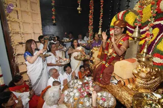 A seated figure in a red-brown dhoti and orange-brown turban sits on a gold-carved throne with a gold lion-like base, wearing a red and white garland, gold necklaces and bangles, and raising both hands in blessing. Around him a crowd of devotees holds silver thalis with lit oil lamps and offerings, many wearing white or red clothes, while a camera operator films at left. Floral garlands hang from the ceiling and brass bells and lamps surround the throne in a dark stone shrine with a large wooden door.