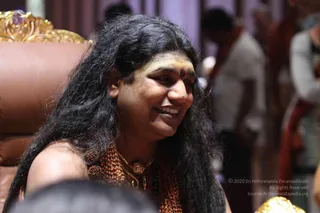 Close photograph of a smiling person with long, dark, wavy hair seated in a brown leather chair with ornate gold trim. The person wears a bare shoulder, a dense rudraksha mala of reddish-brown beads, and a gold pendant necklace, with a yellow-ochre forehead mark and a small red bindi. Blurred people and draped fabric stand in the background of a hall.